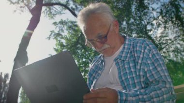 Gözlüklü yaşlı adam parkta bilgisayar kullanmaya çalışıyor.