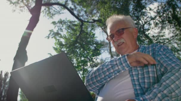 Alter Mann mit Brille beim Versuch, Computer im Park zu benutzen, aber er kippt. Er ruft jemanden an, der ihm helfen kann — Stockvideo