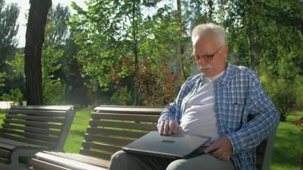En äldre man sitter på bänken med en bärbar dator i trädgården. — Stockvideo