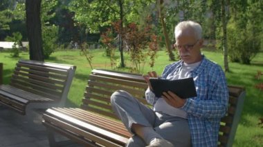 Gözlüklü yaşlı bir adam parkta bir bankta e-kitap okuyor.
