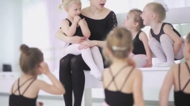 Genç bale öğretmeni, bale okulunda siyah taytlı küçük kızlarla oturuyor ve onlarla iletişim kuruyor.