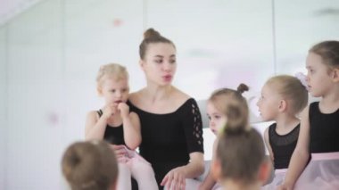 Genç bale öğretmeni, bale okulunda siyah taytlı küçük kızlarla oturuyor ve onlarla iletişim kuruyor.