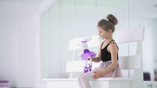 Menina com brinquedo na mão estão sentados no banco em sala de aula de balé — Vídeo de Stock