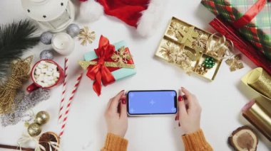 Chroma anahtarı olan akıllı telefon kullanan bir kadın, tıklatıyor, kaydırıyor, kaydırıyor. Beyaz masa arkasında Noel tatili süslemesi.