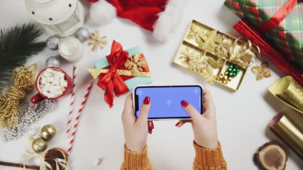 Frau bedient Smartphone mit Chroma-Taste, tippt, wischt, blättert hoch. Weihnachten Feiertagsdekoration auf weißem Tischhintergrund. — Stockvideo