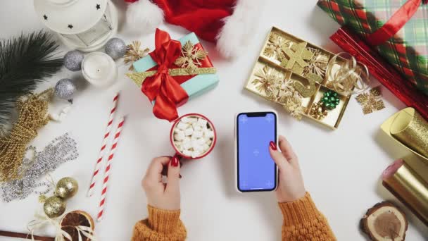 Femme buvant du cacao et utilisant un smartphone avec clé Chroma, tapotant, faisant défiler vers le haut. Décoration de Noël sur fond de table blanche . — Video