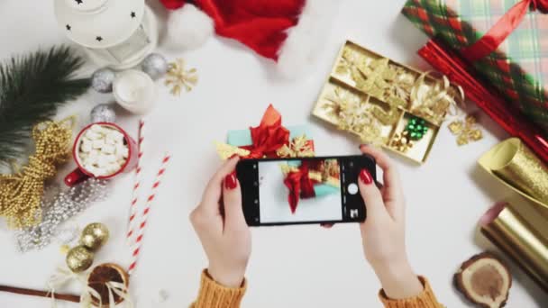 Kvinnan tar en vacker bild på smarttelefonen. Kvinnliga händer photogtaphing julklapp på vitt bord. — Stockvideo