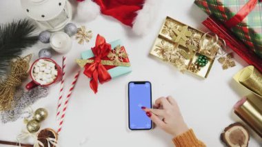 Chroma anahtarı olan akıllı telefon kullanan bir kadın, tıklatıyor, kaydırıyor, kaydırıyor. Beyaz masa arkasında Noel tatili süslemesi.