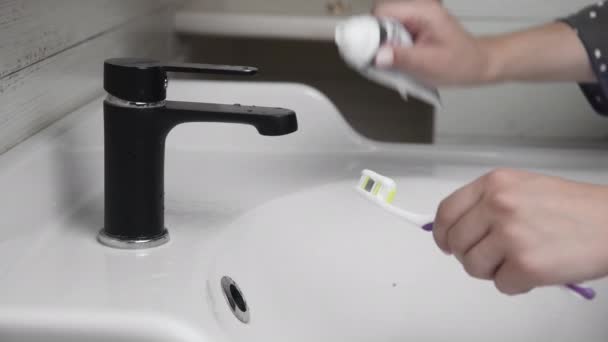 Jonge vrouw poetst tanden met een tandenborstel in de badkamer. Close-up van handen knijpen pasta op de borstel — Stockvideo