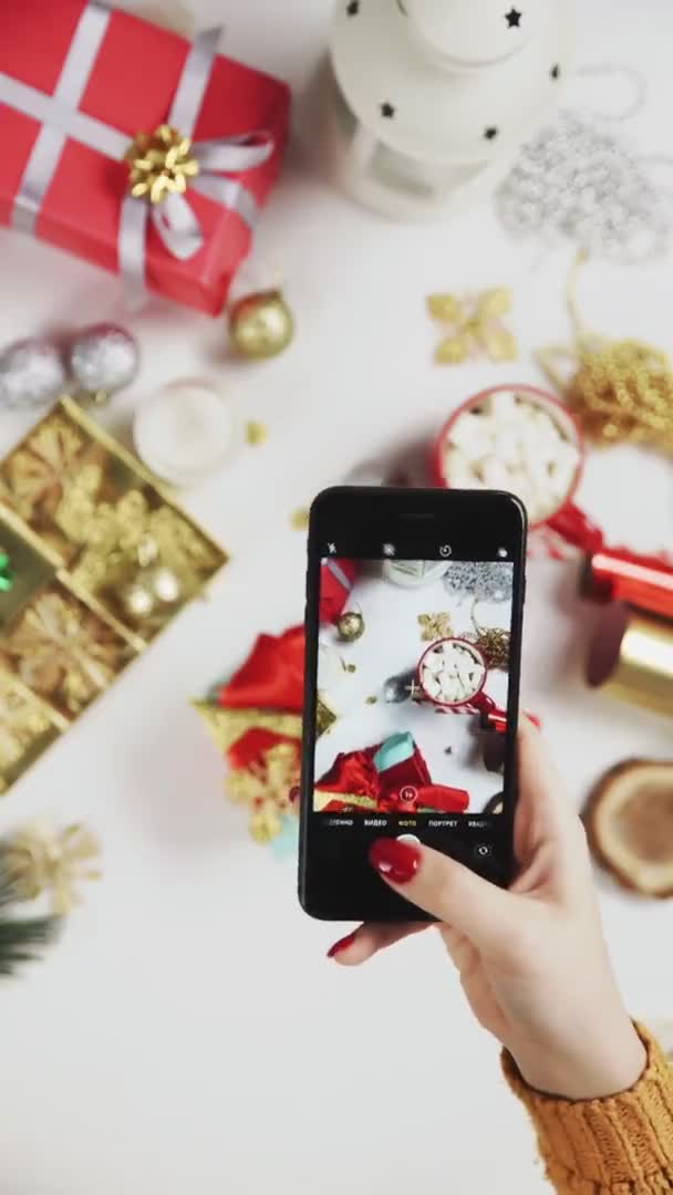 Mujer tomando una hermosa foto en el teléfono inteligente. Manos femeninas photogtaphing regalo de Navidad en la mesa blanca. Vídeo de orientación vertical de la pantalla 9: 16 — Vídeos de Stock