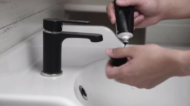 Мужчина сжимает черную гель скраб или черную маску в открытой ладони, стоя в ванной — стоковое видео