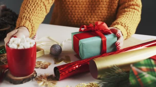 Chica joven bebiendo chocolate caliente con regalos de Navidad en la mesa — Vídeo de stock