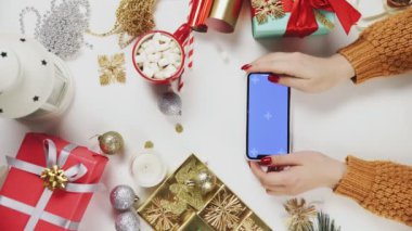 Chroma anahtarı olan akıllı telefon kullanan bir kadın, tıklatıyor, kaydırıyor, kaydırıyor. Beyaz masa arka planında Noel tatili süsü. Dikey Ekran Yönlendirme Videosu 9: 16