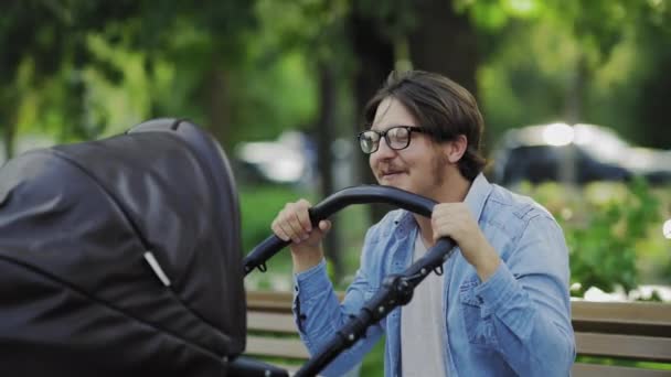 Усміхнений батько розмахує коляскою і розмовляє зі своєю дитиною, відпочиває в парку — стокове відео