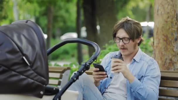 Малыш спит в коляске, отец пользуется сотовым телефоном и пьет кофе . — стоковое видео