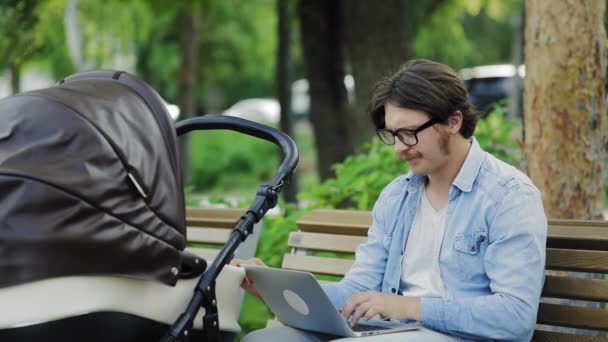 Jovem balançando carrinho de bebê, trabalhando no laptop no parque da cidade, freelancer — Vídeo de Stock