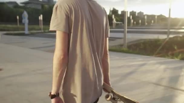 Skateboarder wandelen met skateboard in park witn zonsondergang op de achtergrond — Stockvideo