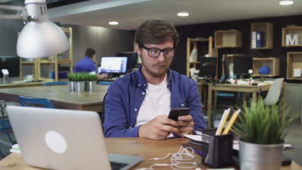 Jeune homme utilise Smartphone alors qu'il est assis au bureau. Homme navigue sur Internet, Montres Vidéos et utilisations Réseaux sociaux — Video