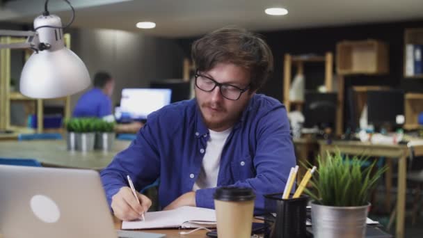 Gözlüklü genç adam ofiste deftere bir şey yazıyor. — Stok video