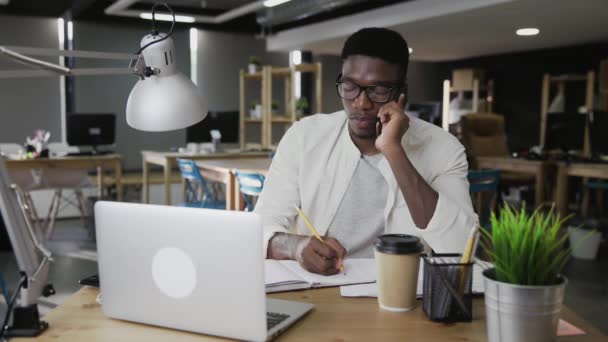 Junger Afrikaner telefoniert mit Handy und macht sich Notizen in modernem Büro — Stockvideo