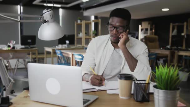 アフリカ人男性は携帯電話で話していると現代のオフィスでメモを取っている — ストック動画