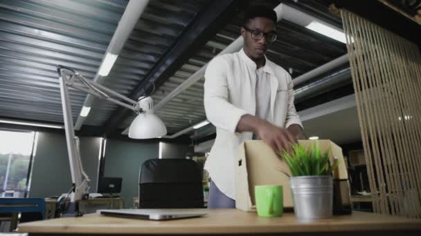 L'uomo africano si trasferisce in un nuovo ufficio. Sta imballando oggetti personali in una scatola di cartone. — Video Stock