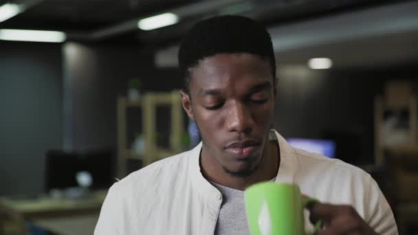 Porträt eines attraktiven schwarzen Mannes mit Smartphone-Gerät, das SMS-Nachrichten verschickt und in einem modernen Büro Kaffee oder Tee trinkt — Stockvideo