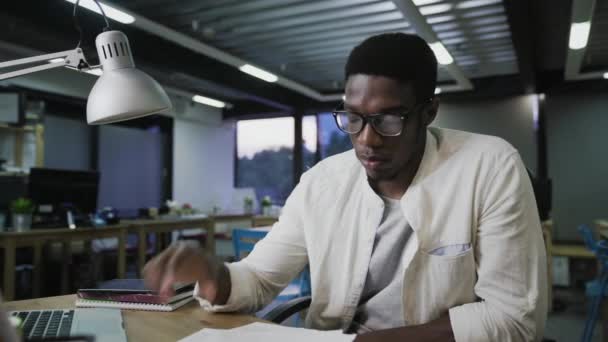 若いアフリカ人男性で眼鏡をかけてオフィスのテーブルに座って書類を読む — ストック動画