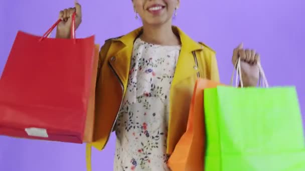 Portret van Afro-Amerikaanse vrouw met boodschappentassen op paarse achtergrond in Studio. Happy Woman Holding Shopping Kleurrijke Tassen. Gele jas — Stockvideo