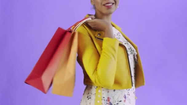 Портрет африканской американки с пакетами покупок на фиолетовом фоне в Студии. Счастливая женщина держит в руках разноцветные сумки. Желтая куртка — стоковое видео