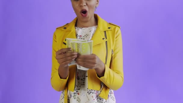 Mulher afro-americana atraente está contando dinheiro contra um fundo roxo jaqueta amarela — Vídeo de Stock