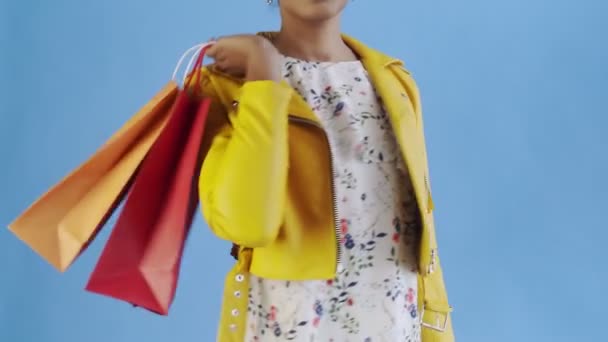 Retrato de mujer afroamericana con bolsas de compras y abanico de dinero sobre fondo azul en Studio. Chaqueta amarilla — Vídeos de Stock