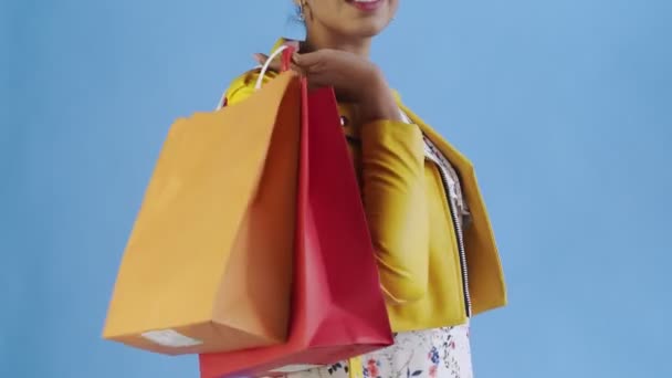 Retrato de mujer afroamericana con bolsas de compras y abanico de dinero sobre fondo azul en Studio. Chaqueta amarilla — Vídeos de Stock