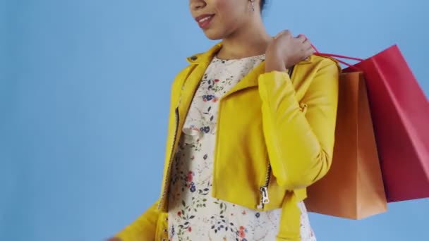 Retrato de mujer afroamericana con bolsas de compras y teléfono móvil sobre fondo azul en Studio. Chaqueta amarilla — Vídeos de Stock