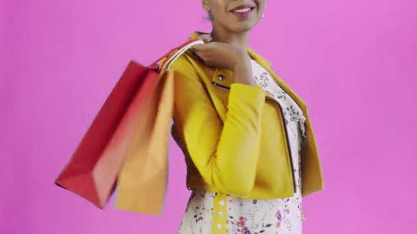 Portré afro-amerikai nő bevásárló táskák rózsaszín háttér stúdióban. Boldog nő kezében vásárlás színes táskák. Sárga kabát — Stock videók