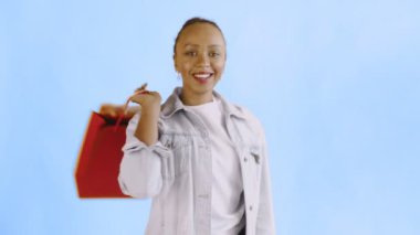Stüdyoda mavi arka planda alışveriş torbaları taşıyan Afrikalı Amerikalı bir kadının portresi. Mutlu Kadın Alışveriş Çantalarını Tutuyor. Kot ceket.