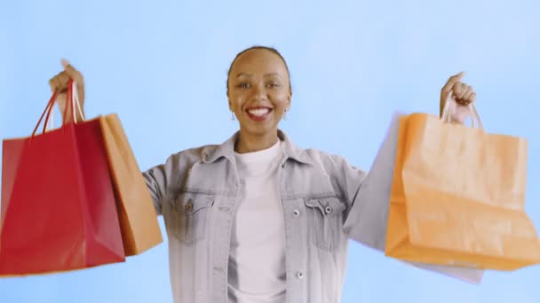 Retrato de mulher afro-americana com sacos de compras em fundo azul no Studio. Happy Woman Holding Sacos coloridos de compras. Casaco Jean — Vídeo de Stock