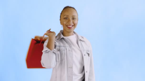 Porträt einer afrikanisch-amerikanischen Frau mit Einkaufstaschen auf blauem Hintergrund im Studio. glückliche Frau mit bunten Einkaufstaschen. Jeansjacke — Stockvideo