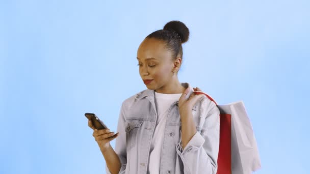 Portret van Afrikaanse Amerikaanse vrouw met boodschappentassen praat met smartphone op blauwe achtergrond in Studio. Jean-jas — Stockvideo
