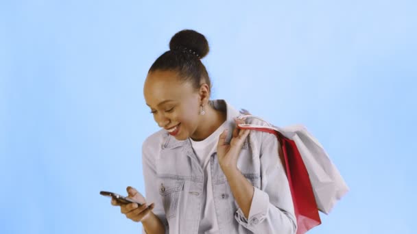 Alışveriş çantalı Afro-Amerikan kadın portresi Stüdyoda mavi arka planda akıllı telefondan konuşuyor. Kot ceket. — Stok video
