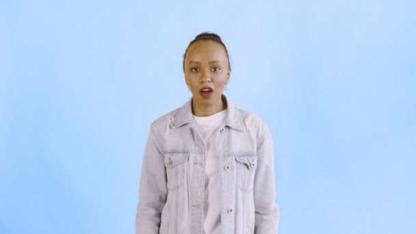 Skrämd afrikansk amerikansk kvinna rädd för något och tittar in i kameran med stora ögon full av skräck över blå bakgrund. Jean jacka — Stockvideo