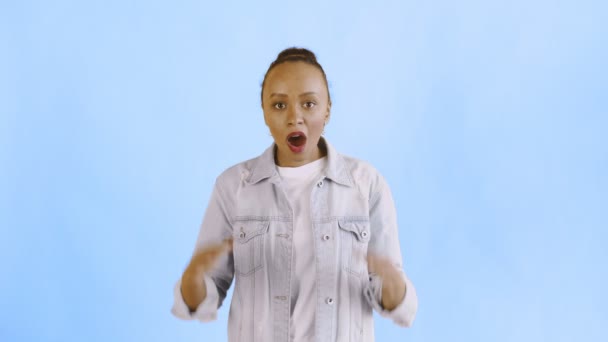 Frightened african american woman looks into the camera with open mouth and lifts hands to head on blue background. Jean jacket — Stock Video