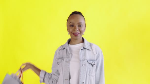 Retrato de mujer afroamericana con bolsas de la compra está mostrando los pulgares hacia arriba en el fondo amarillo en el estudio. Chaqueta Jean — Vídeo de stock