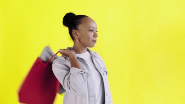 Retrato de una mujer afroamericana con bolsas de compras está bebiendo un café sobre fondo amarillo en Studio. Chaqueta Jean — Vídeos de Stock