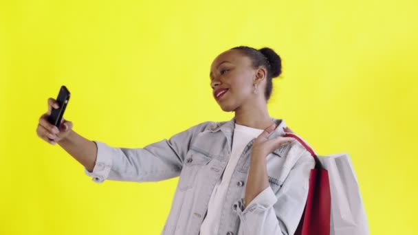 Retrato de una mujer afroamericana con bolsas de compras está haciendo una selfie sobre fondo amarillo en Studio. Chaqueta Jean — Vídeo de stock