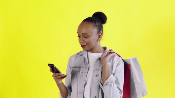 Retrato de mulher afro-americana com sacos de compras está falando por smartphone no fundo amarelo no Studio. Casaco Jean — Vídeo de Stock