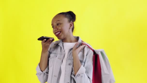 Retrato de mujer afroamericana con bolsas de la compra está grabando un mensaje de voz por teléfono inteligente en fondo amarillo en el estudio. Chaqueta Jean — Vídeo de stock