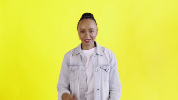 Skott av ung afrikansk amerikansk kvinna ler glatt med armarna korsade tittar på kameran på gul bakgrund Jean jacka — Stockvideo