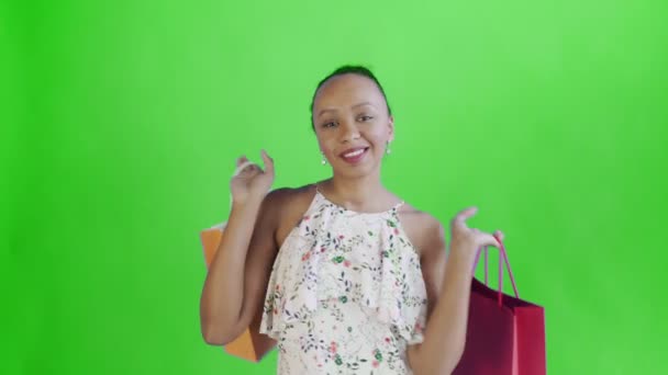 Mulher afro-americana está posando com sacos de compras em ambas as mãos no fundo verde no Studio. Happy Woman Holding Sacos coloridos de compras. Vestido branco com flores — Vídeo de Stock