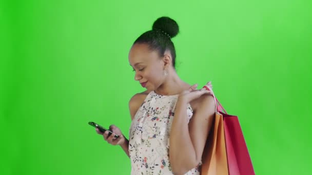 Портрет афро-американської жінки з сумками для покупок розмовляє смартфоном на зеленому фоні в студії. Біла сукня з квітами. — стокове відео